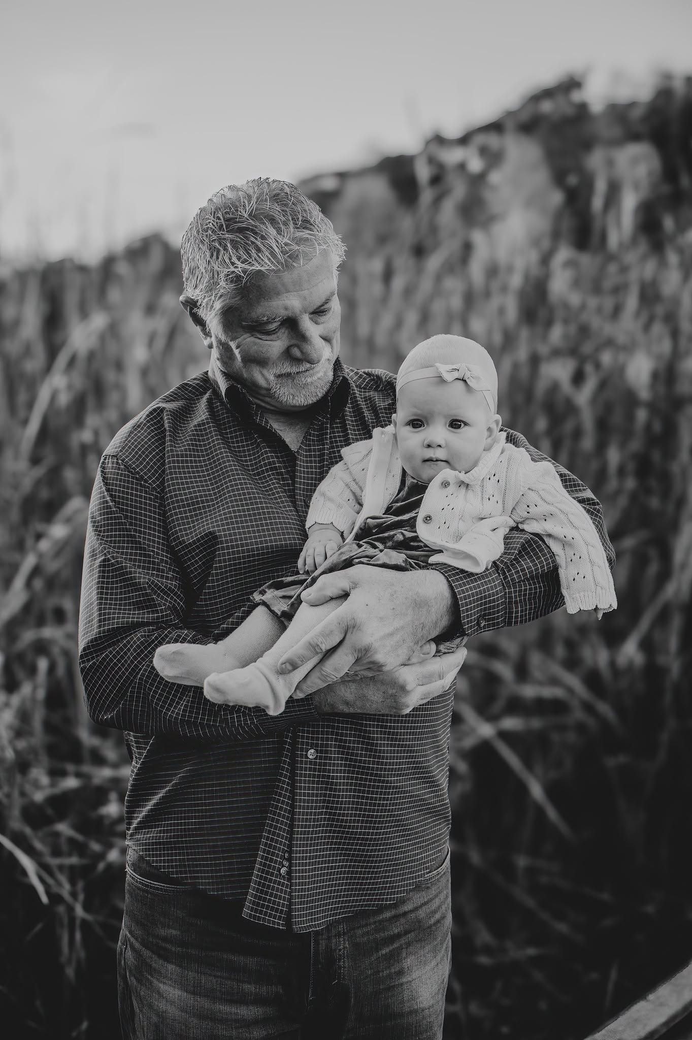 Black and white photo of a grandfather tenderly holding a baby girl outdoors near tall grass.