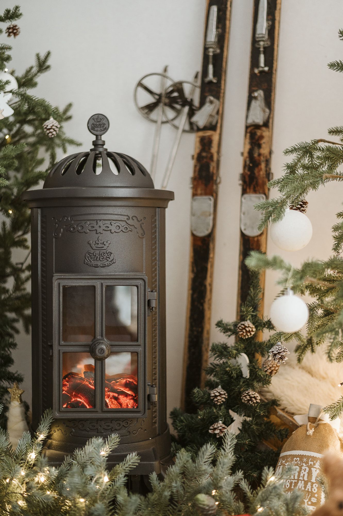 Vintage cast iron stove with glowing faux fire beside vintage wooden skis and a pinecone-decorated Christmas tree with white ornaments.