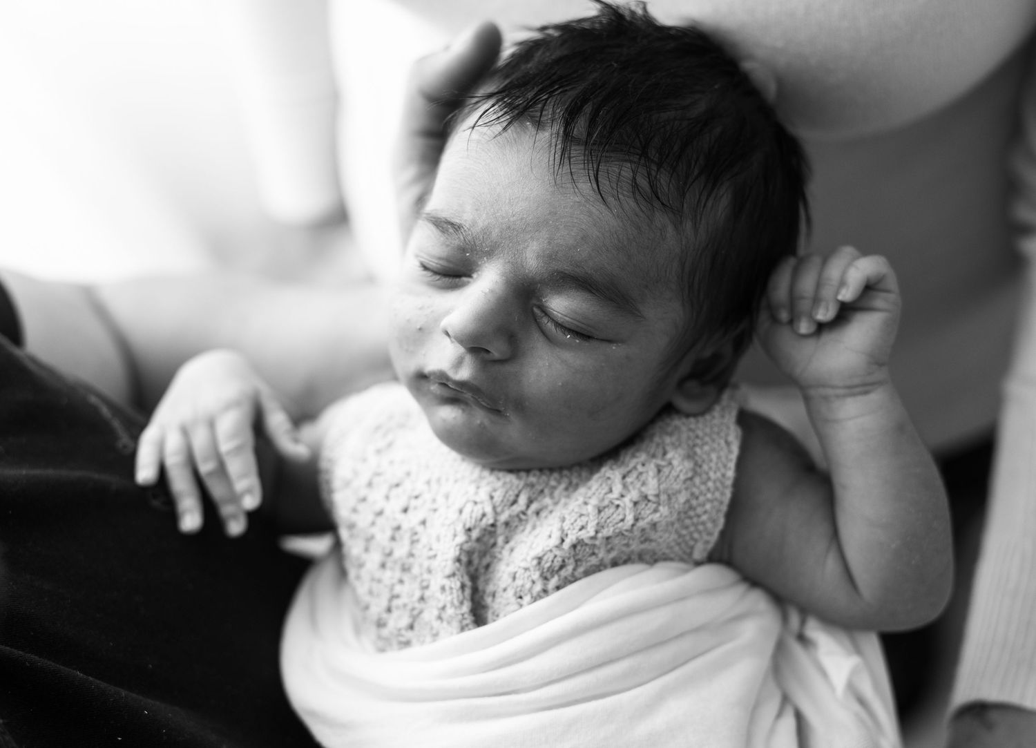 A peaceful sleeping newborn baby stretches while wearing a white knit outfit in this black and white photograph.