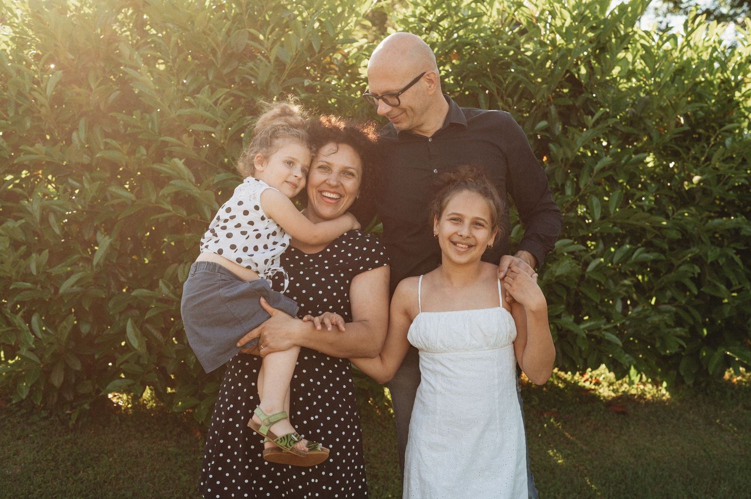 A happy family shares a candid moment together outdoors in warm sunlight against green foliage.