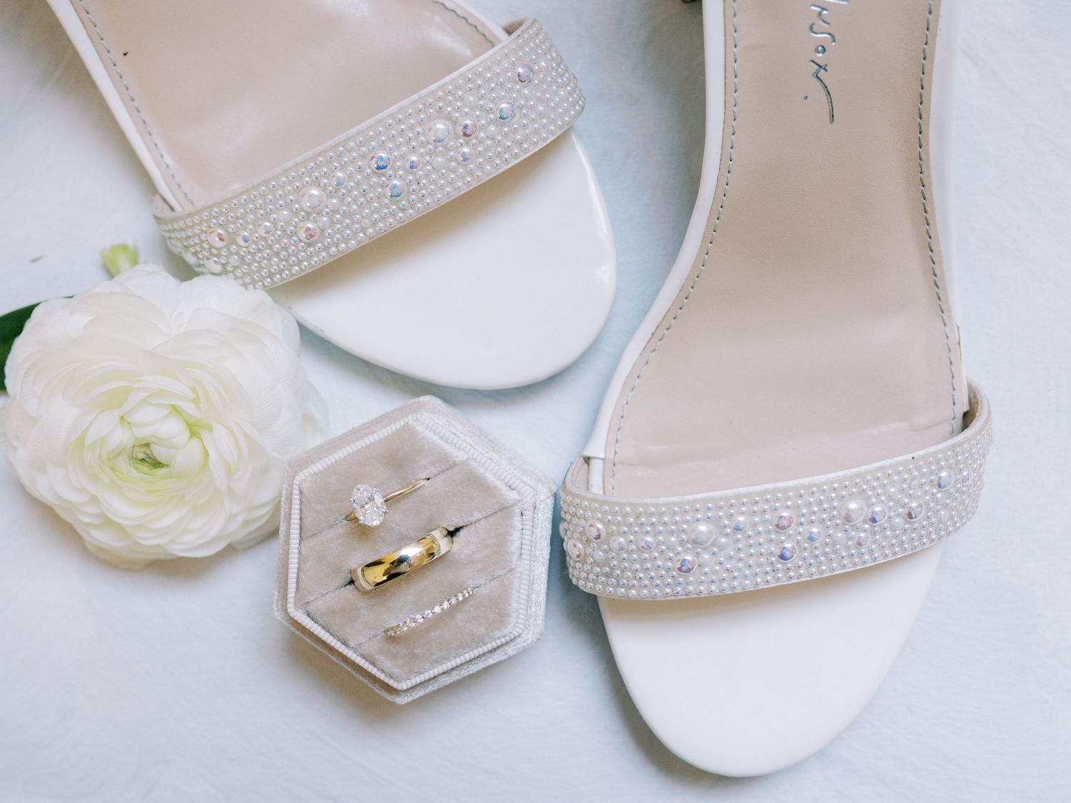 Elegant white bridal sandals with rhinestone details next to hexagonal ring box.