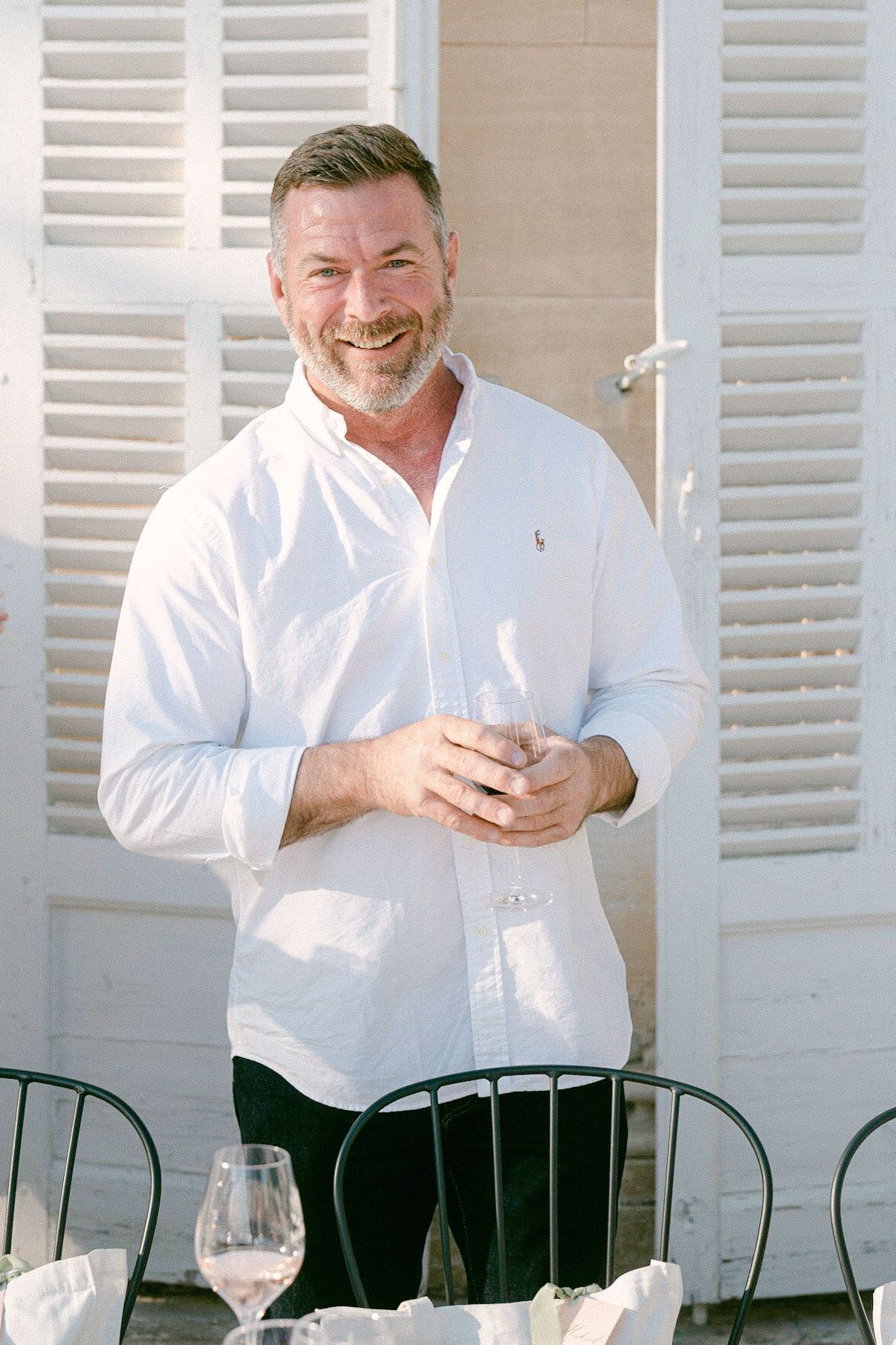 A person in white clothing smiles cheerfully while standing near a dining setup with white shutters.