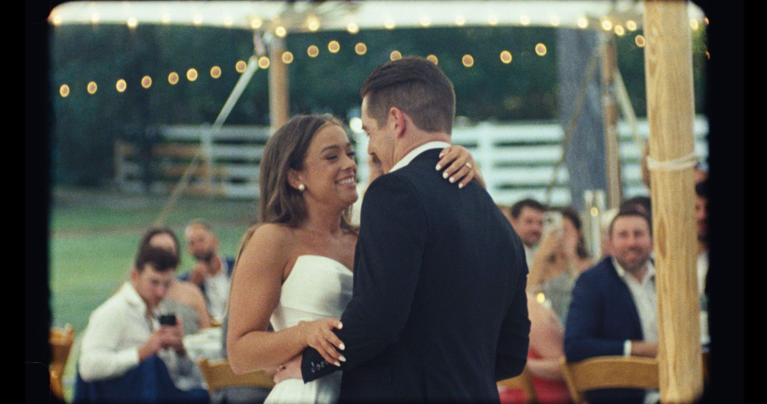A couple shares an intimate dance moment under string lights at an outdoor wedding reception.