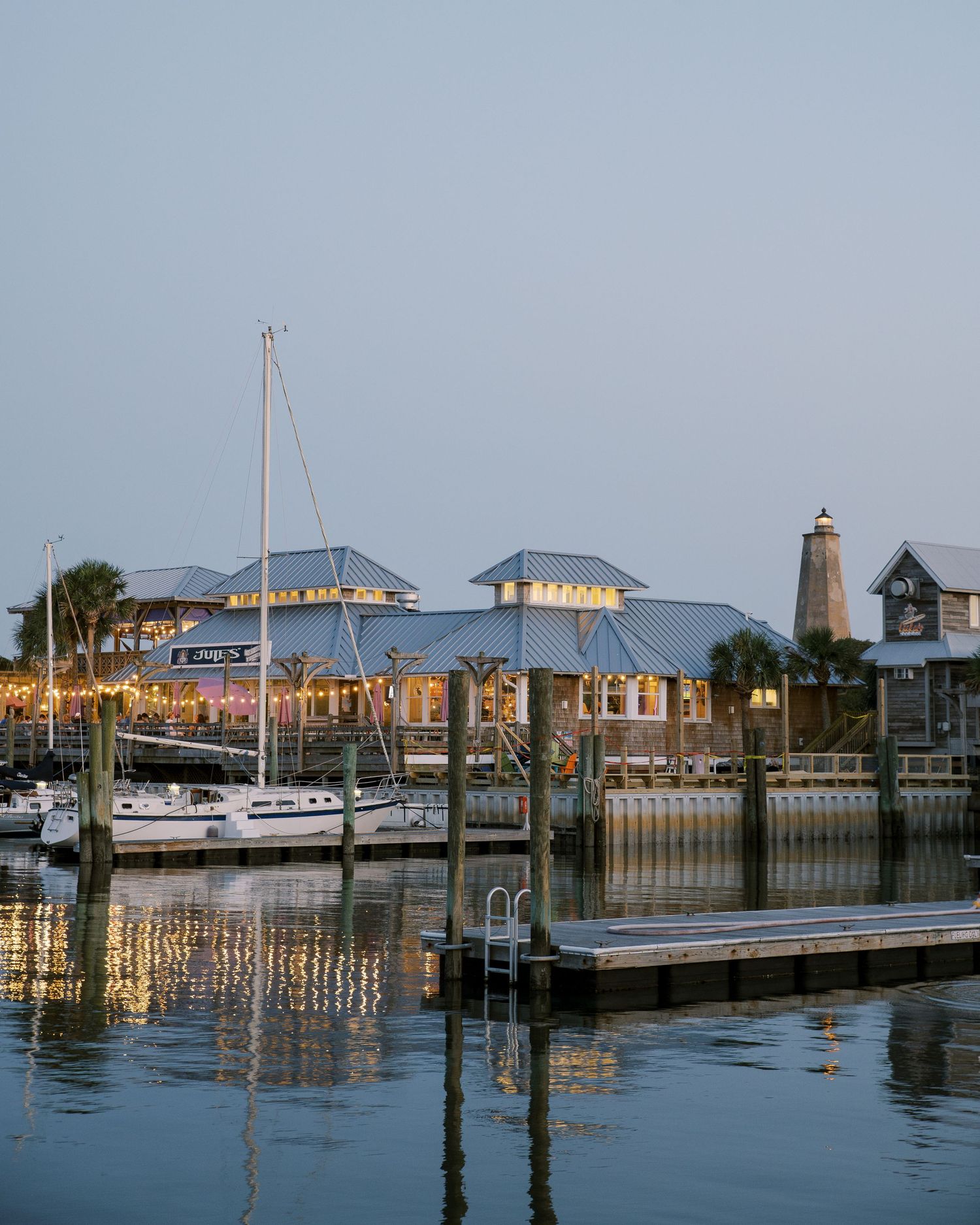 A coastal marina with boats and lighthouse at dusk featuring blue-roofed buildings.