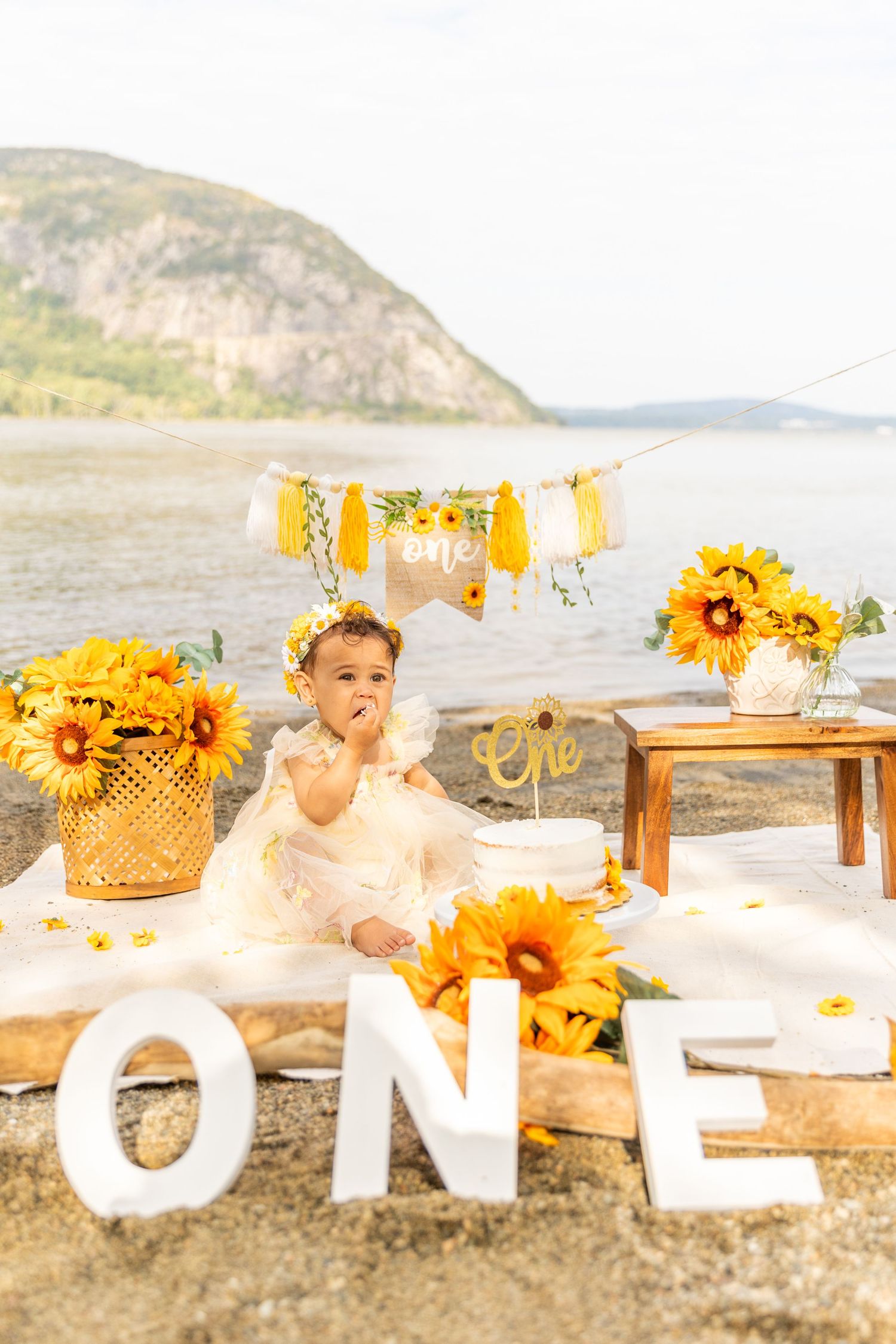 Baby girl in white dress eating cake at sunflower-themed outdoor first birthday ONE celebration.