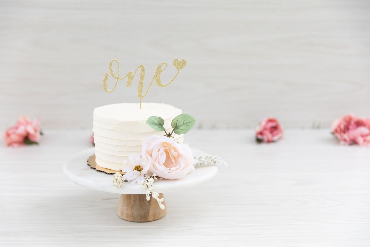 Elegant first birthday smash cake with gold one topper and pink roses on white cake stand.