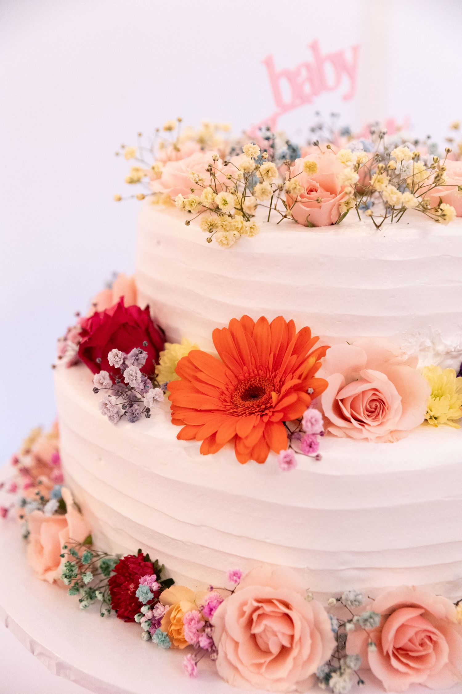 Two-tier white baby shower cake decorated with colorful fresh flowers and a pink baby topper.