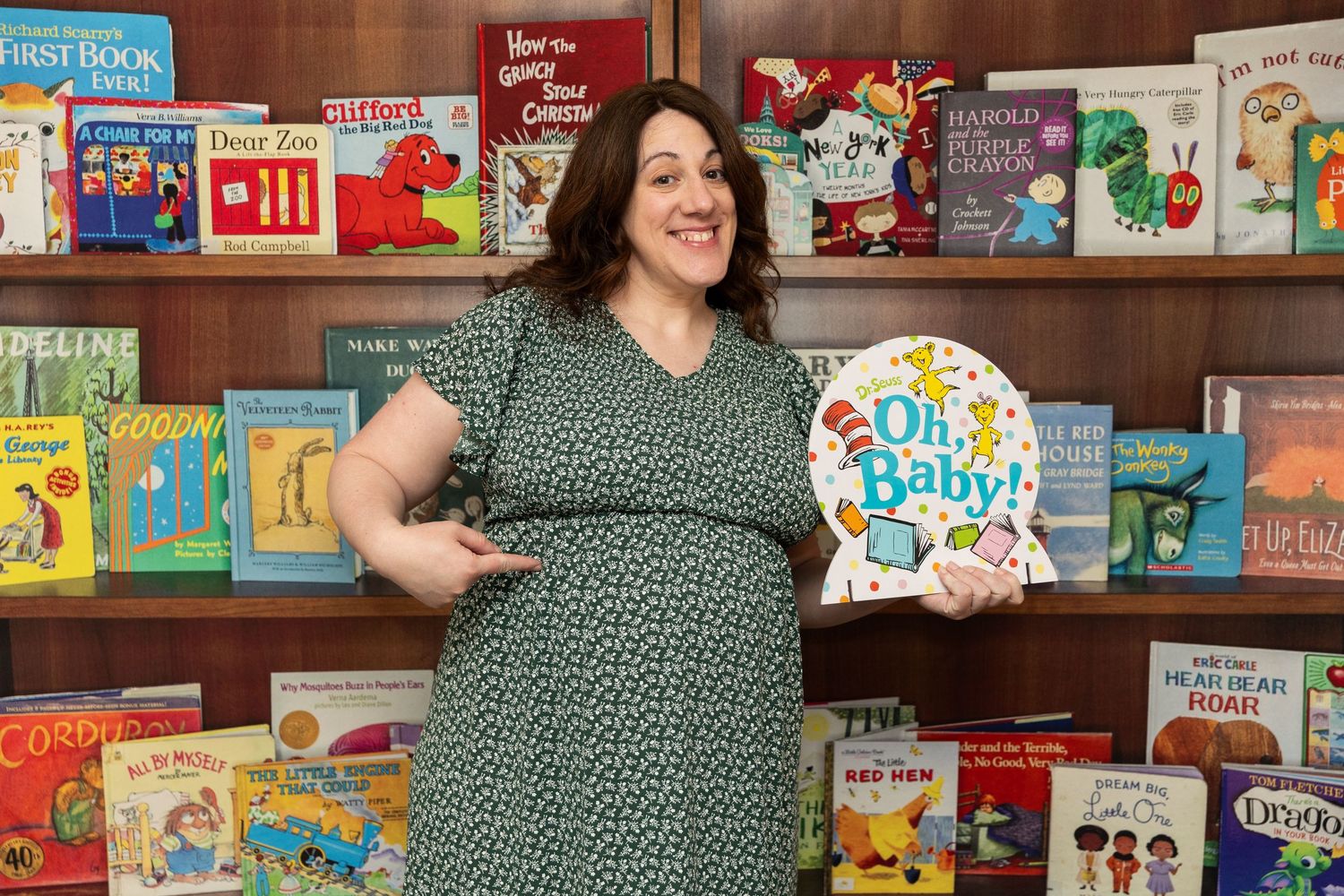 Pregnant woman in green floral dress holding Oh Baby sign at a book-themed baby shower.