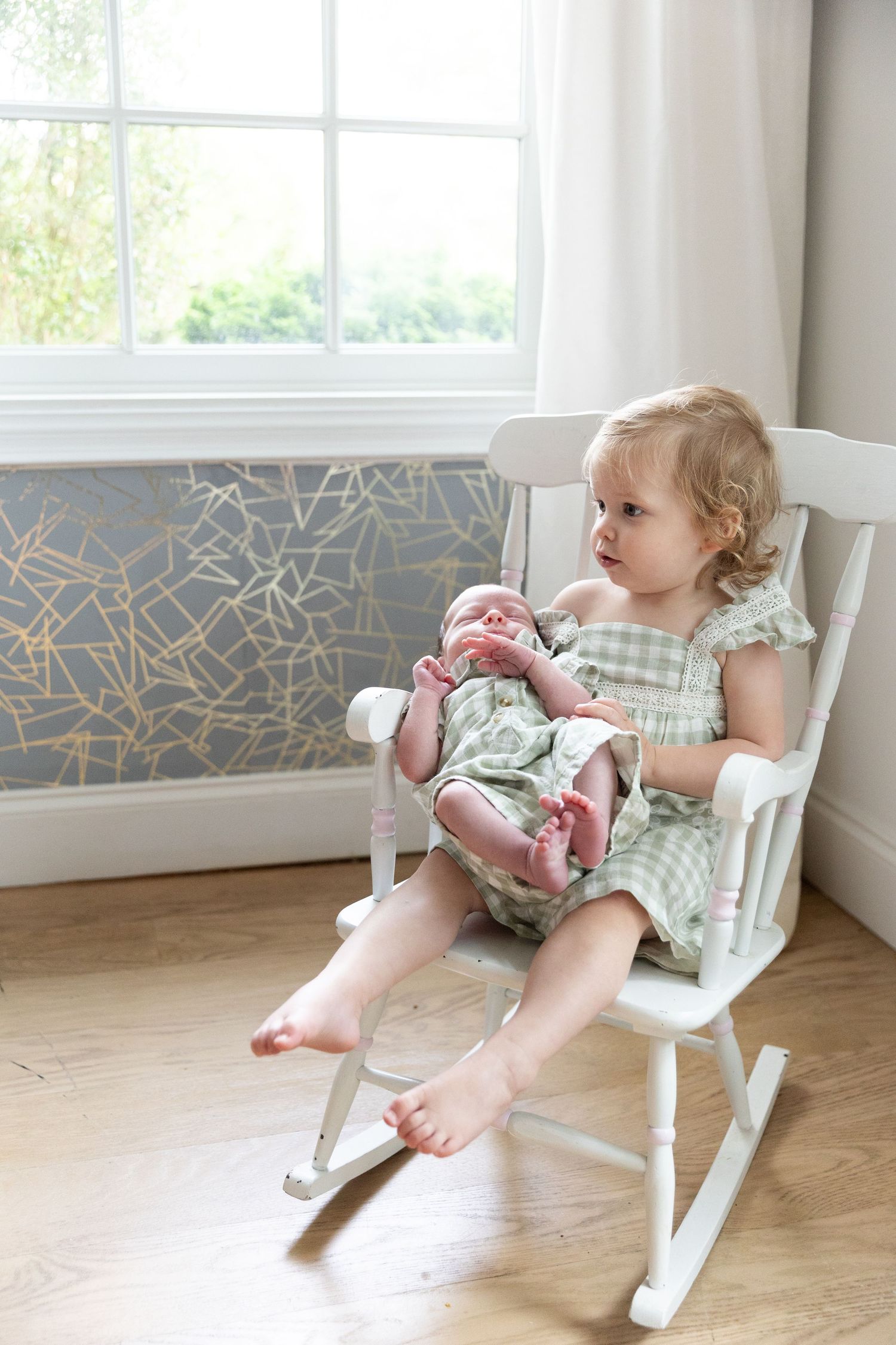 Toddler girl in green dress sitting in white rocking chair holding sleeping newborn baby.