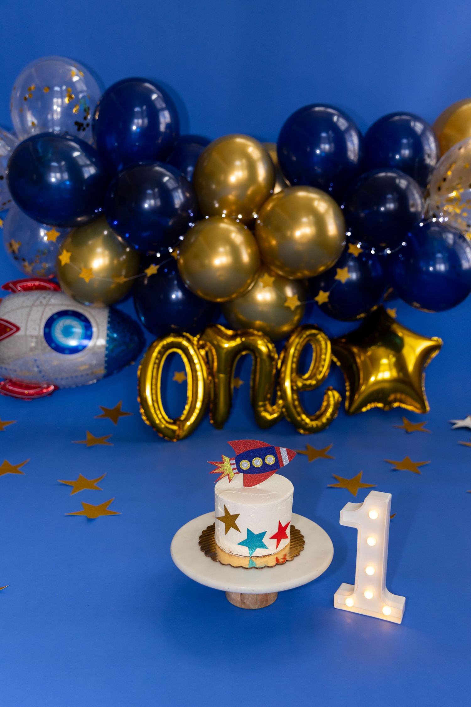 Space themed first birthday setup with rocket cake, gold one balloons, and blue balloon arch.