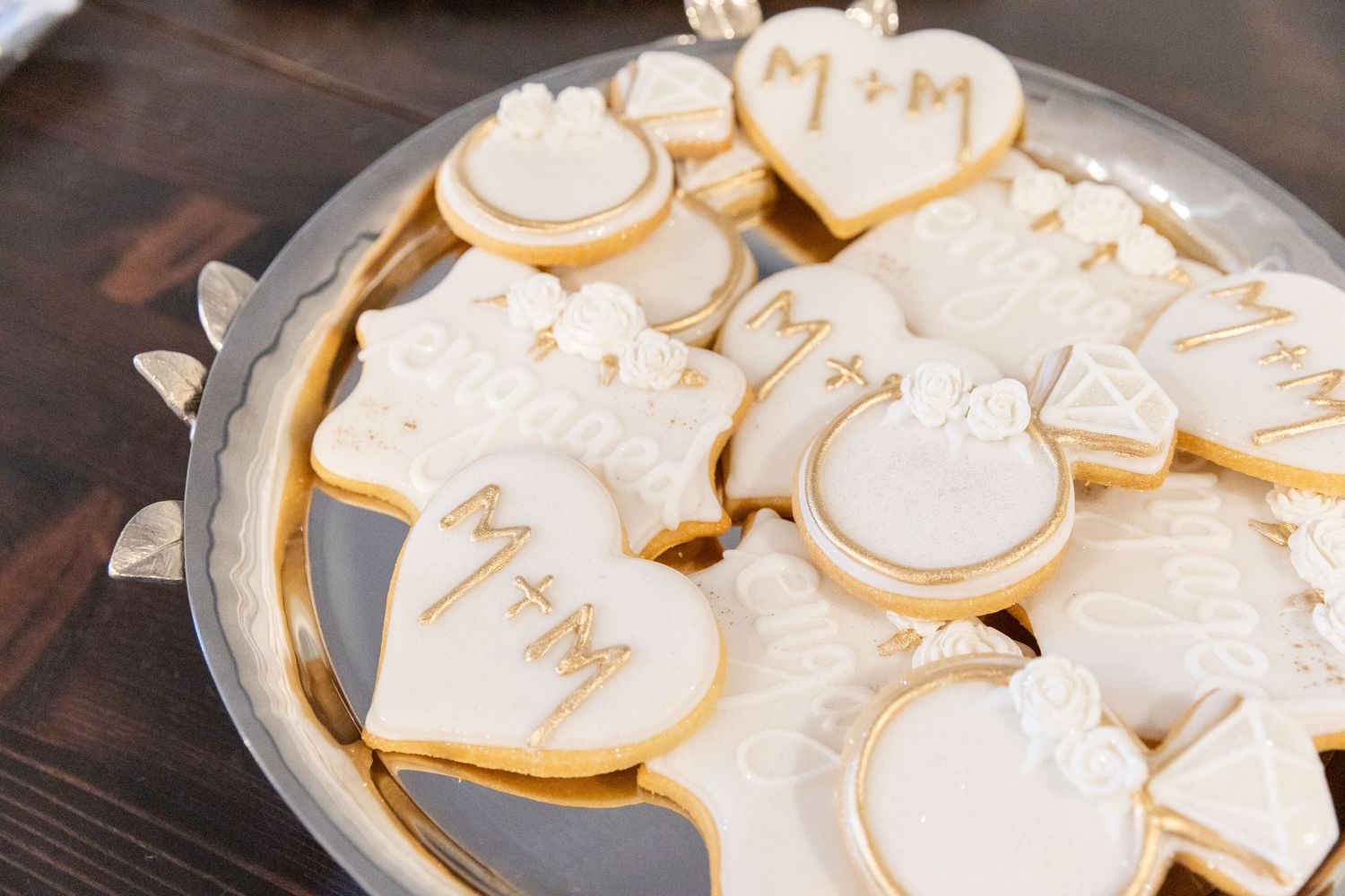 Decorated white and gold sugar cookies with wedding motifs including rings and initials on a silver tray.