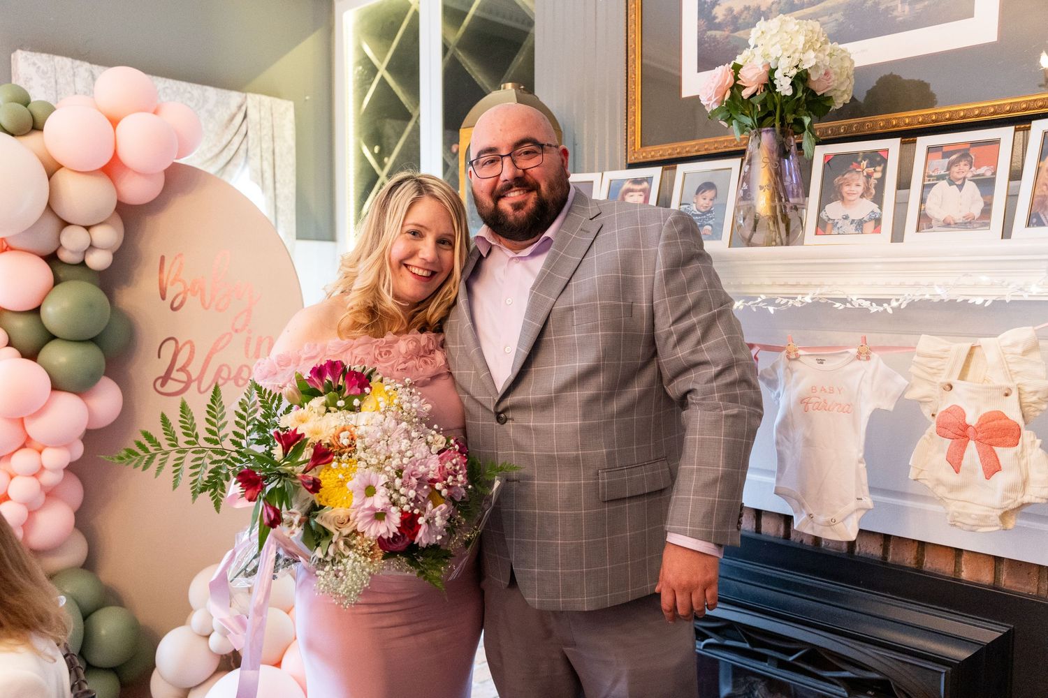Smiling couple posing together at baby shower with balloon garland and framed photos in background.