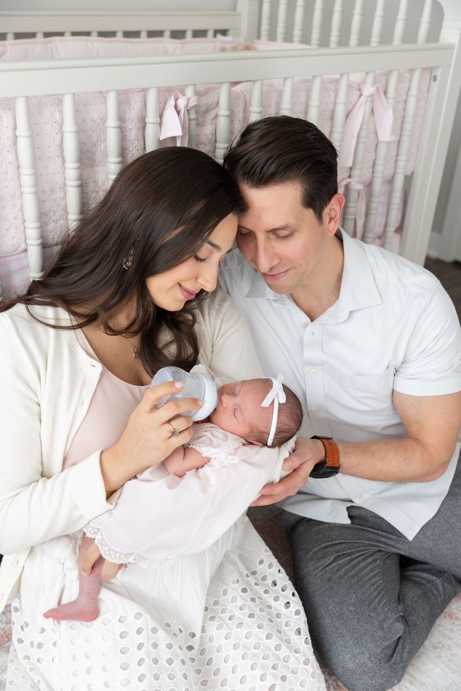 New parents bottle feed their newborn baby girl in a pink nursery beside a white crib.