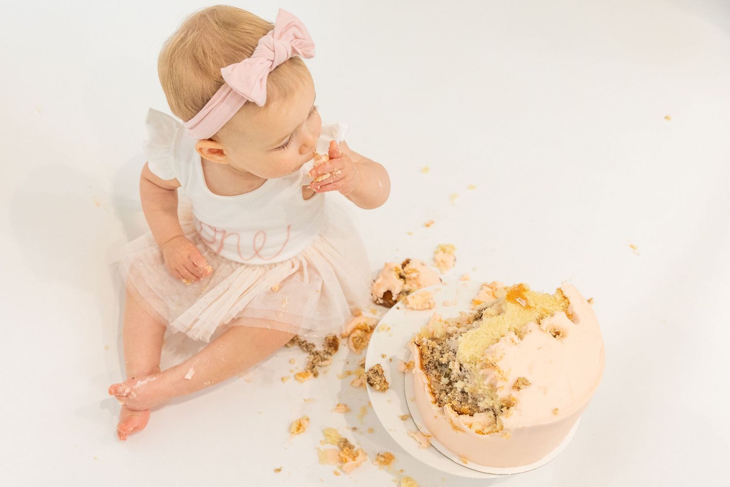 Baby girl in white tutu smashing first birthday cake from above on white background.