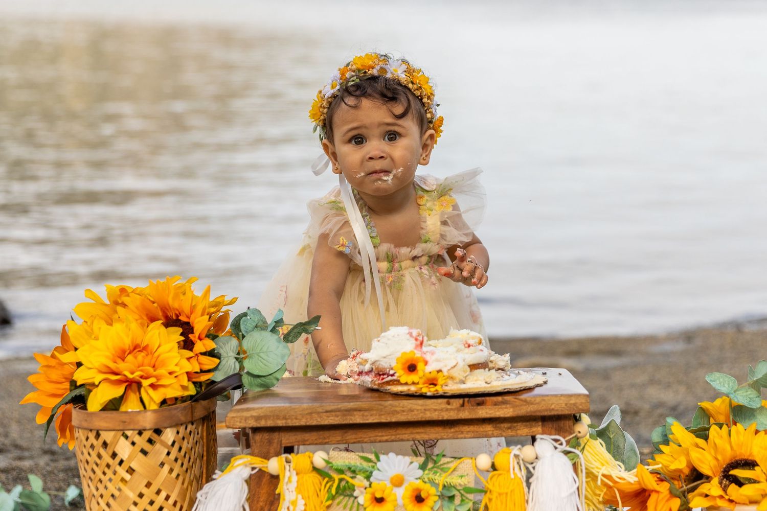 Baby girl with sunflower crown standing at wooden table with cake at outdoor birthday celebration.