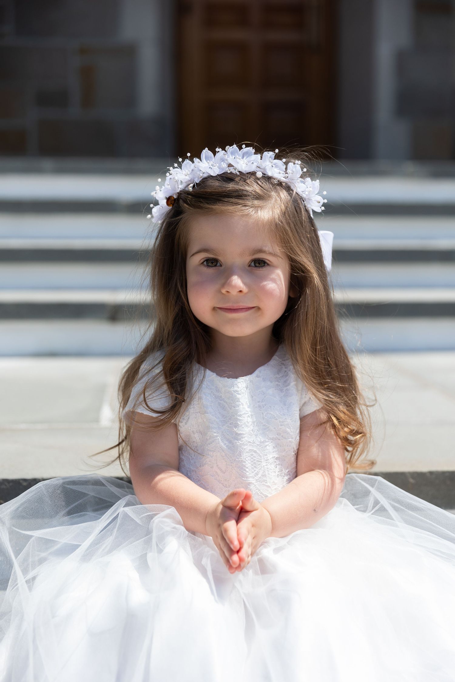 Adorable little girl in white dress and floral crown sitting on church steps.
