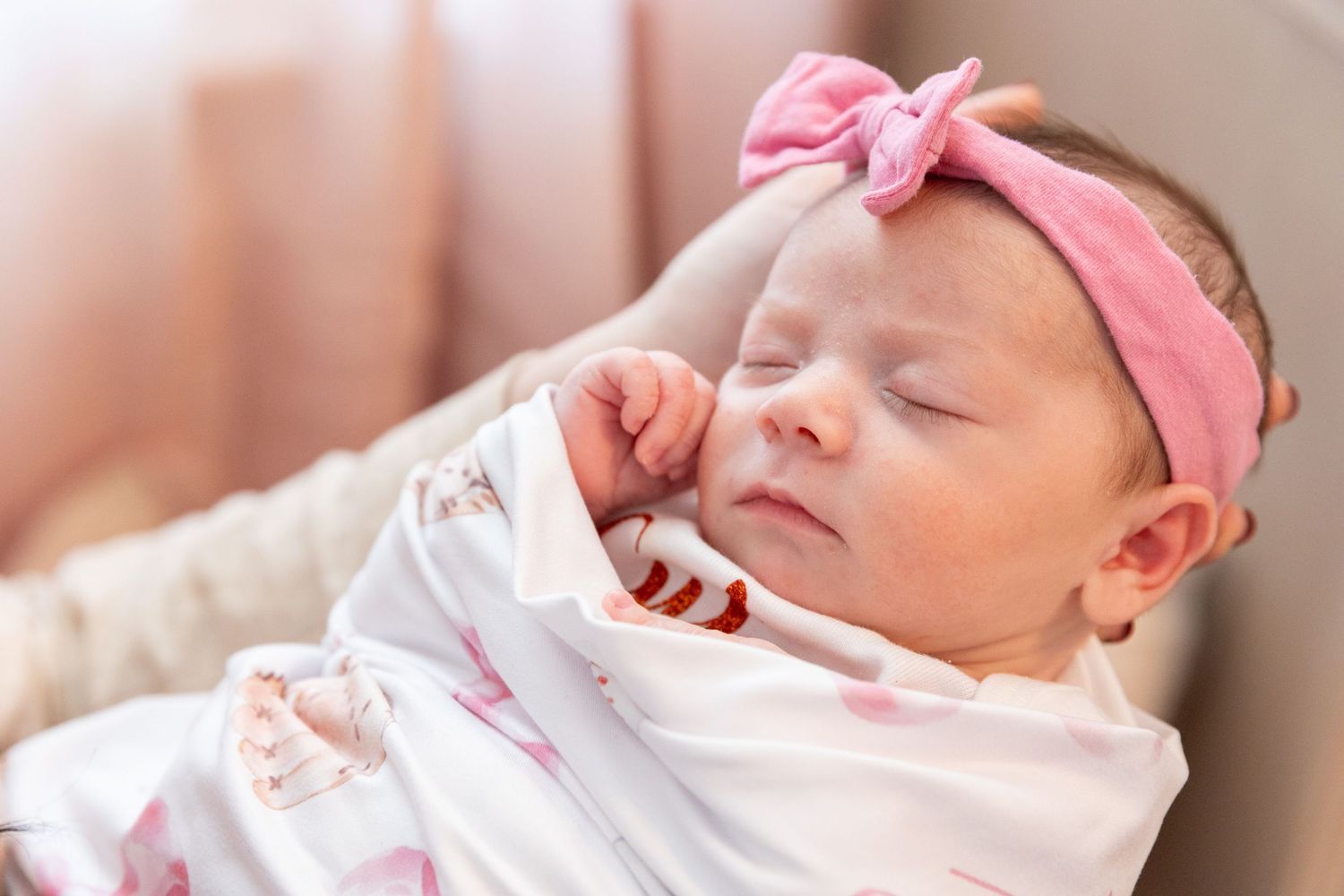 Sleeping newborn baby girl with pink bow headband swaddled in a floral blanket in a bassinet.