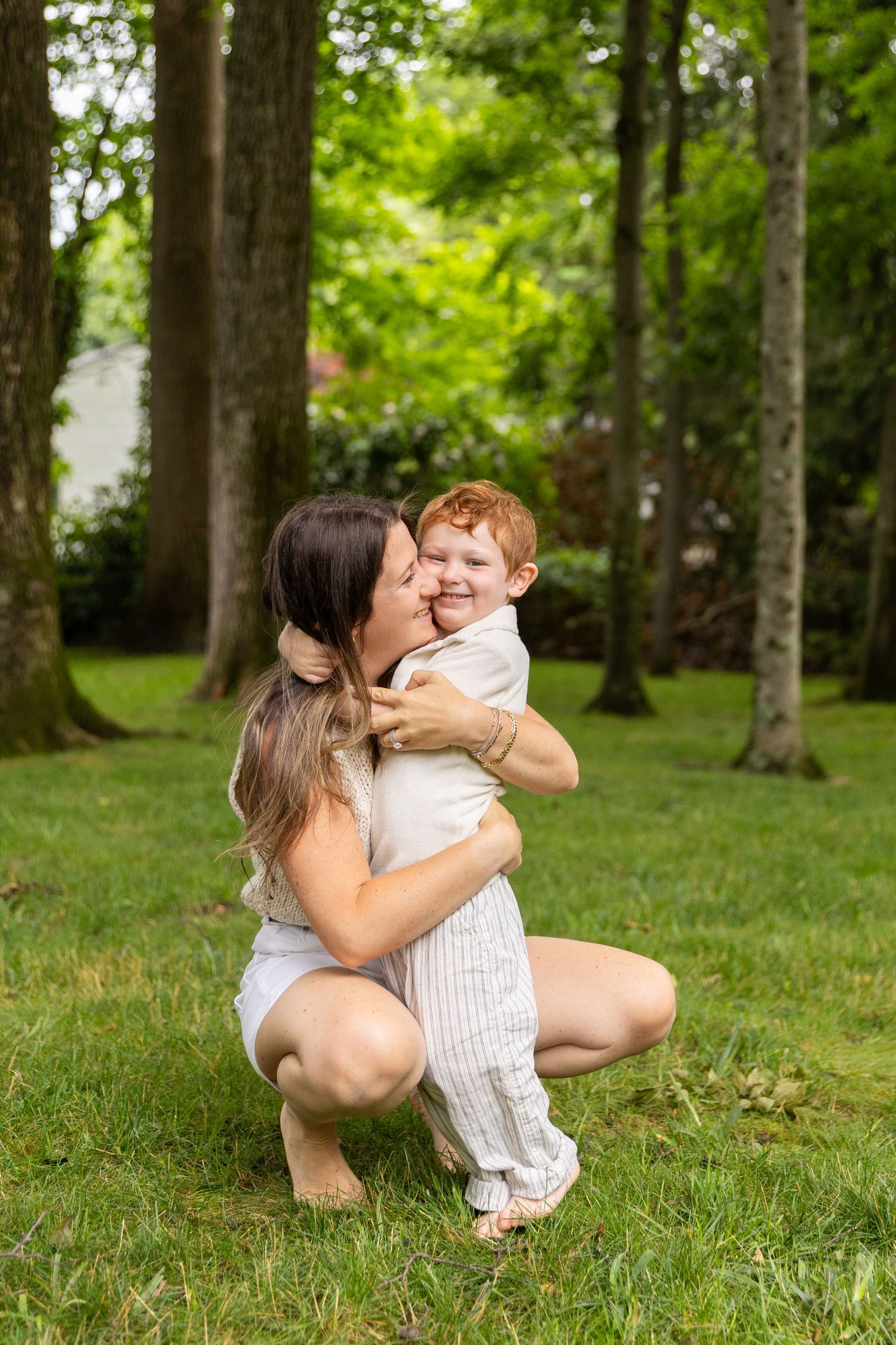 Mother crouching in park hugging smiling red-haired toddler boy among tall trees on green grass.