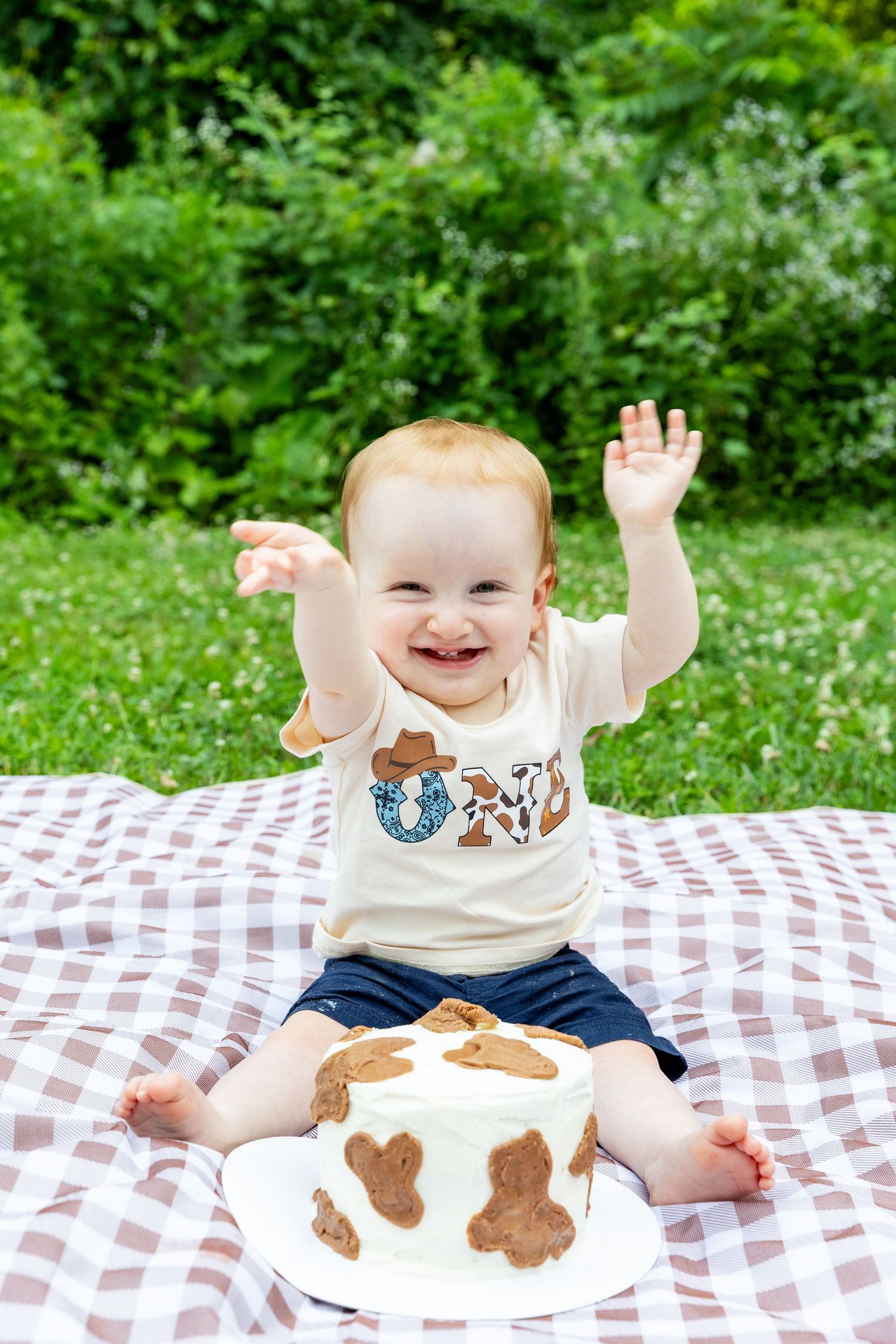Laughing baby raising hands beside cowboy-themed first birthday cake on outdoor picnic blanket.