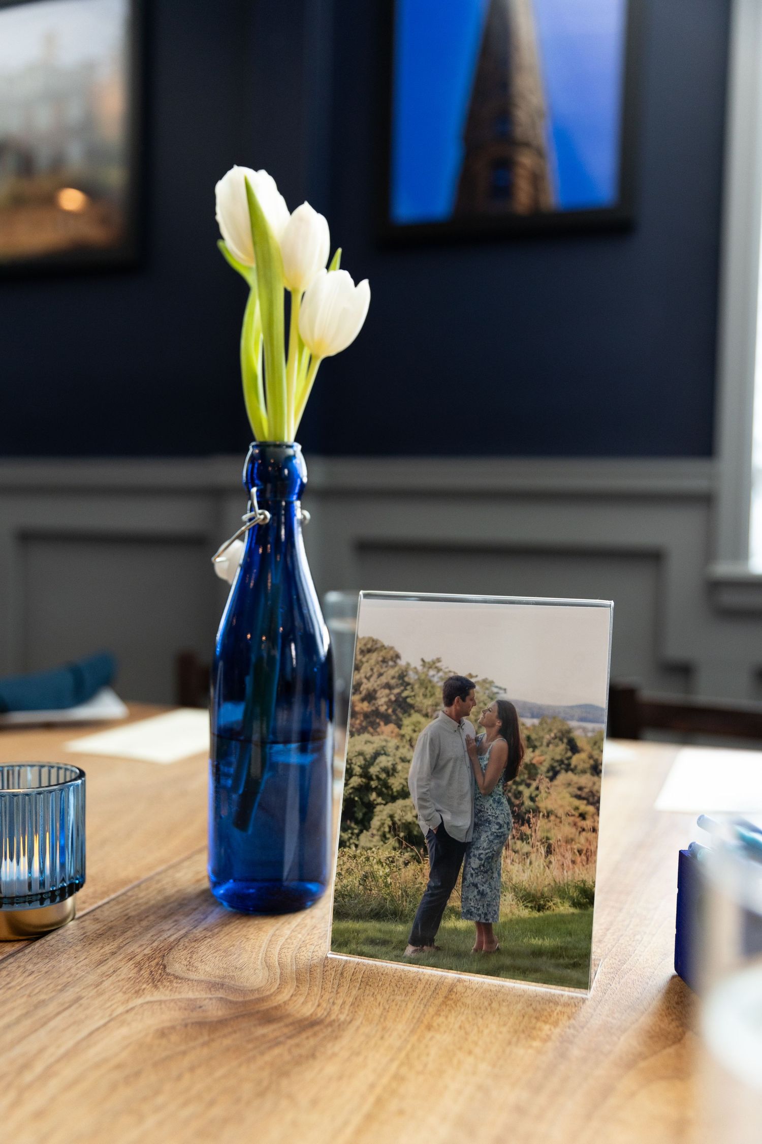 Blue glass vase with white tulips beside a couple's photo frame on wooden table.
