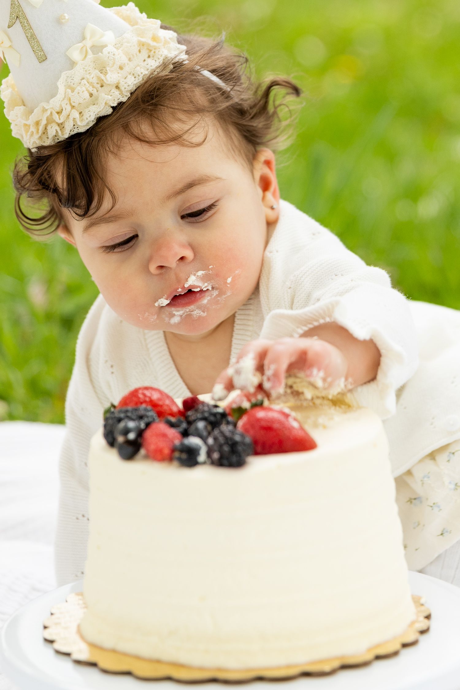 Curly-haired baby in white outfit with party hat eating white berry-topped smash cake outdoors.