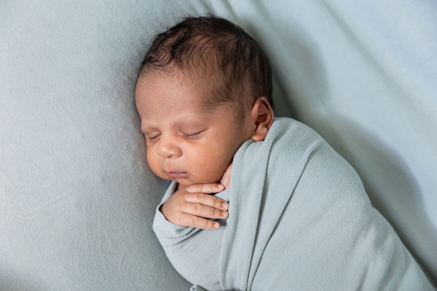 Sleeping newborn baby swaddled in a soft blue blanket with tiny hands tucked under chin.