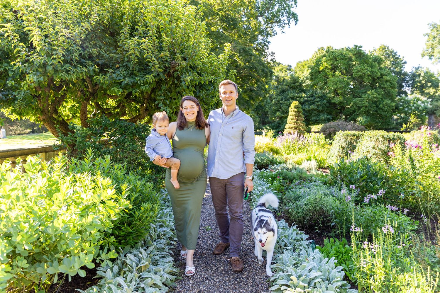 Pregnant couple with toddler and dog posing in lush garden path during summer family maternity session.