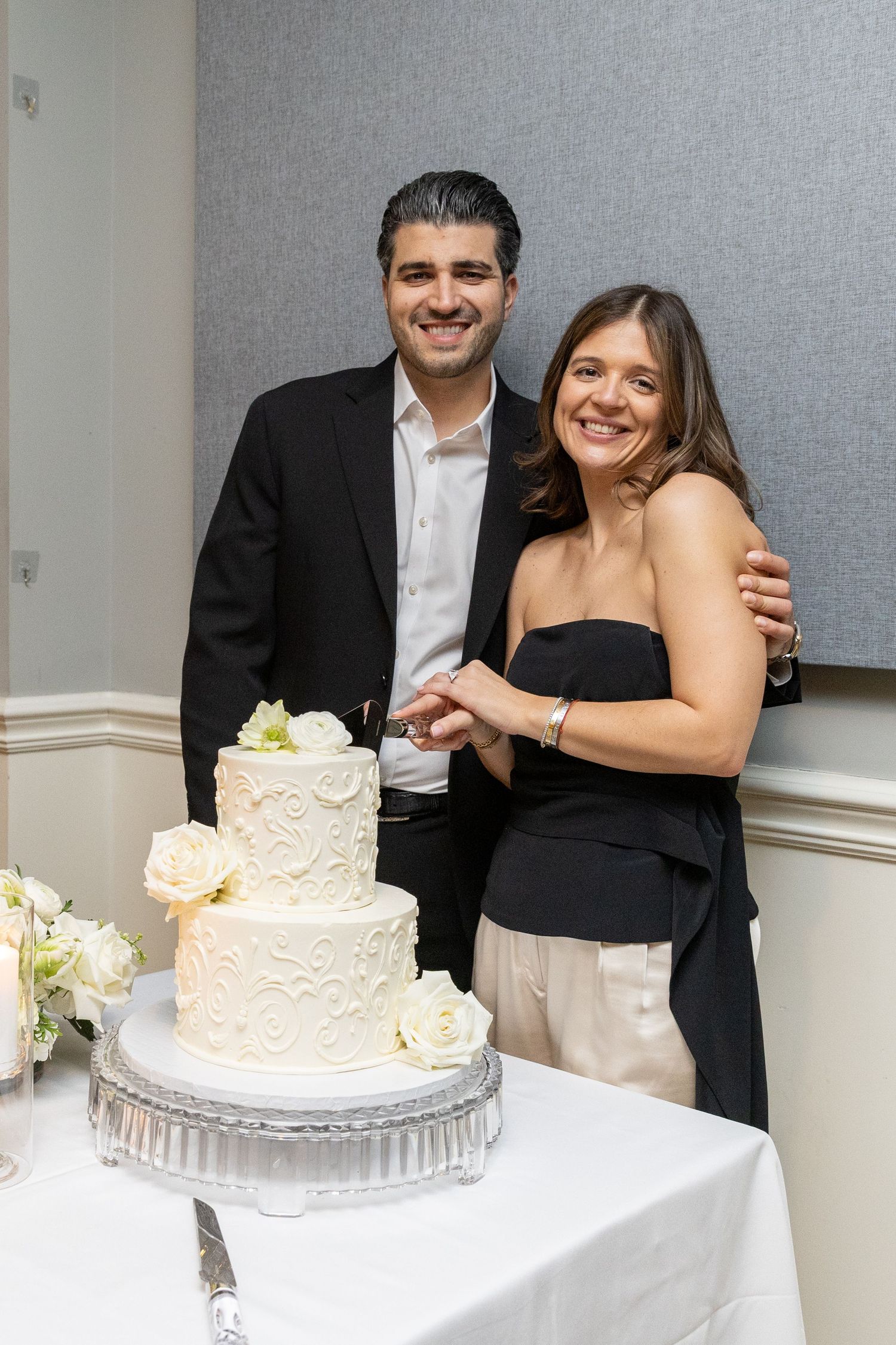 Smiling couple cutting a white three-tier cake decorated with floral frosting at their reception.