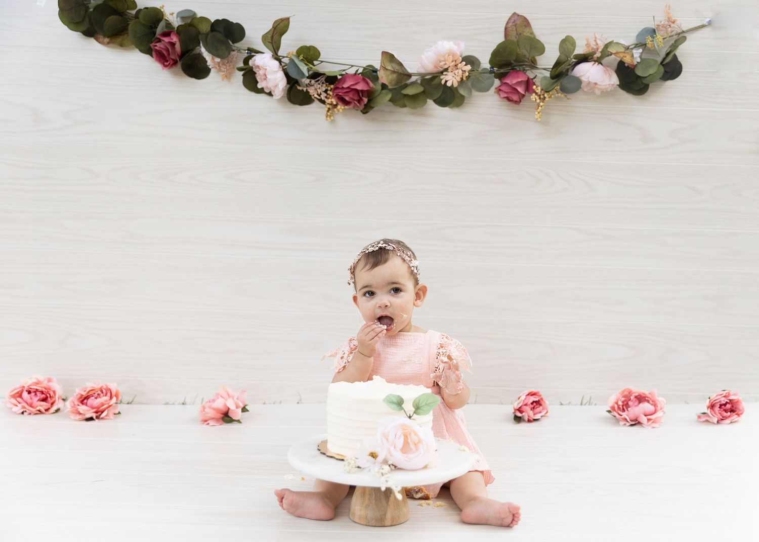 Baby girl in pink dress smashing white birthday cake with floral garland backdrop and roses.