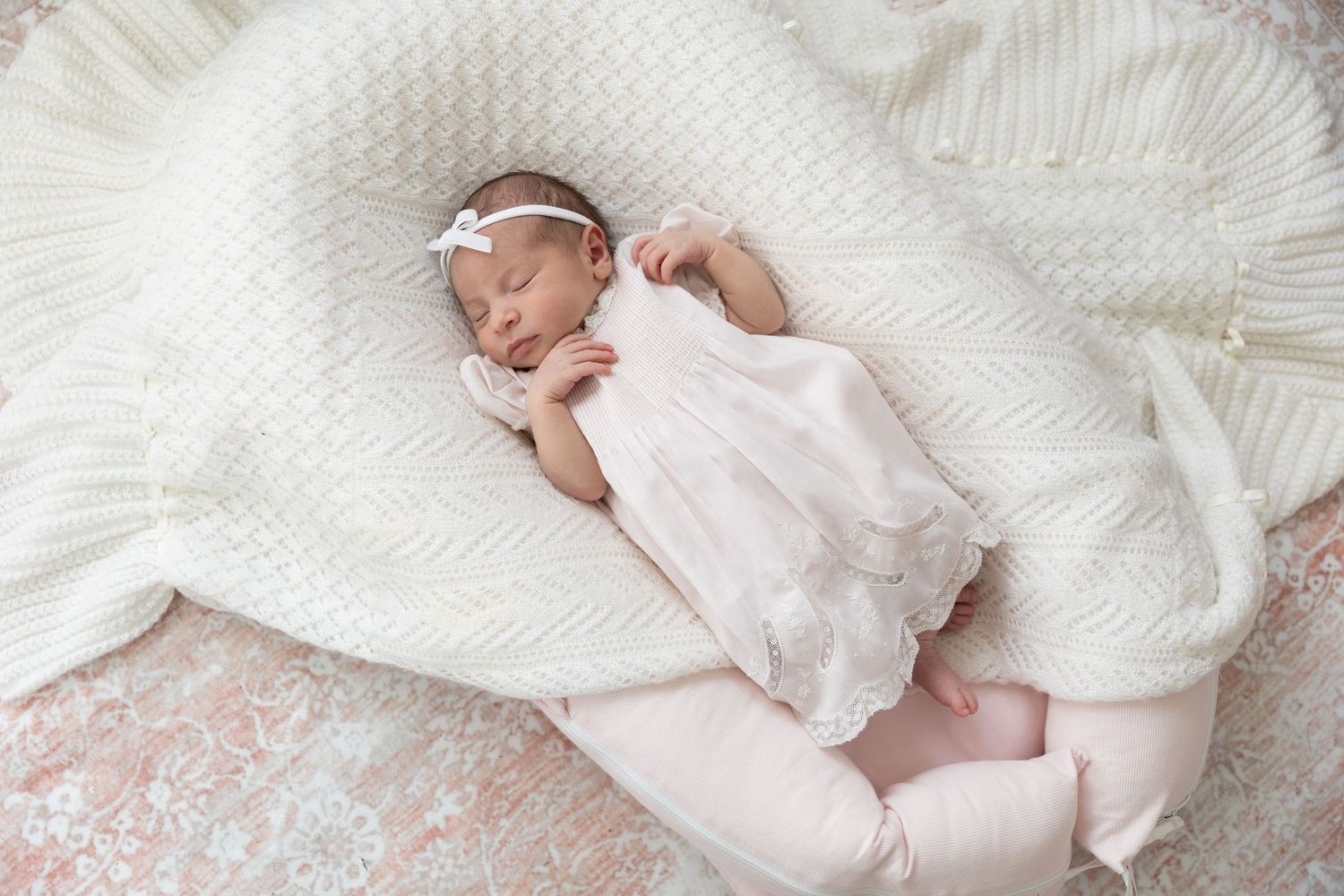 Peaceful newborn baby girl in white lace outfit and floral headband sleeps on a soft white blanket.