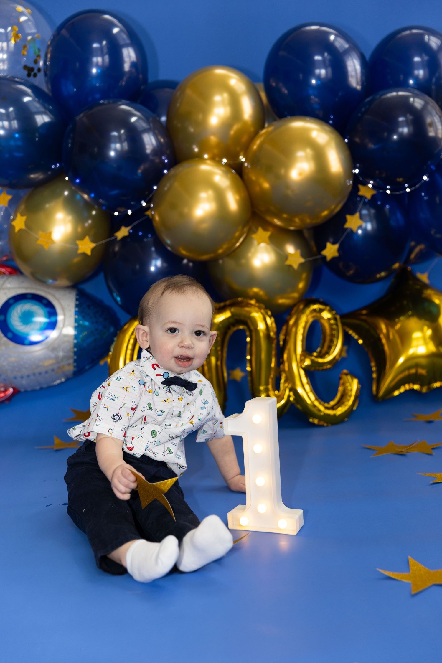 Baby boy in bow tie sitting with number one light up sign and blue gold balloons.