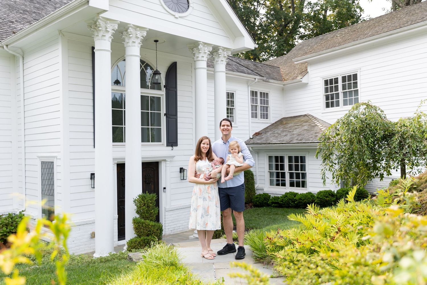 Happy young family with newborn baby posing outside a beautiful white colonial home.