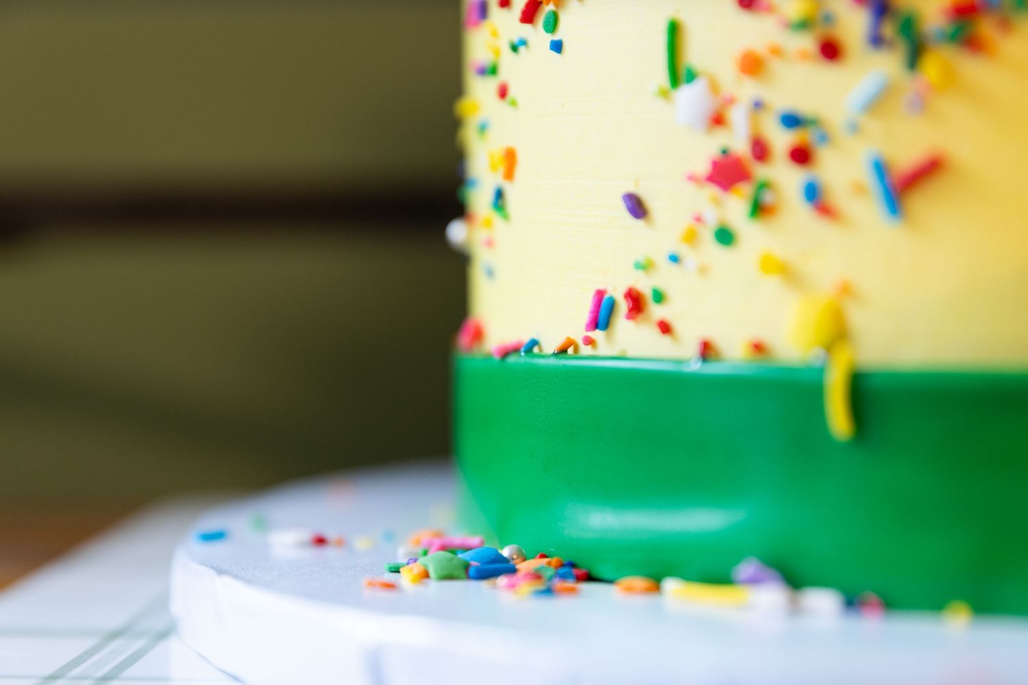 Close-up of colorful sprinkle-covered yellow and green birthday cake on white cake stand.