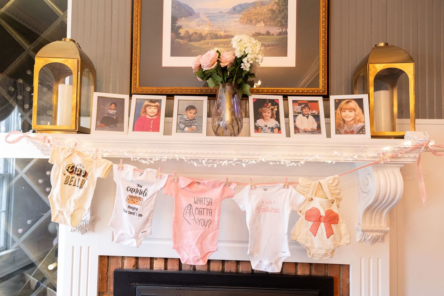 Baby shower fireplace mantel with onesie banner, framed photos, gold lanterns, and floral arrangement.