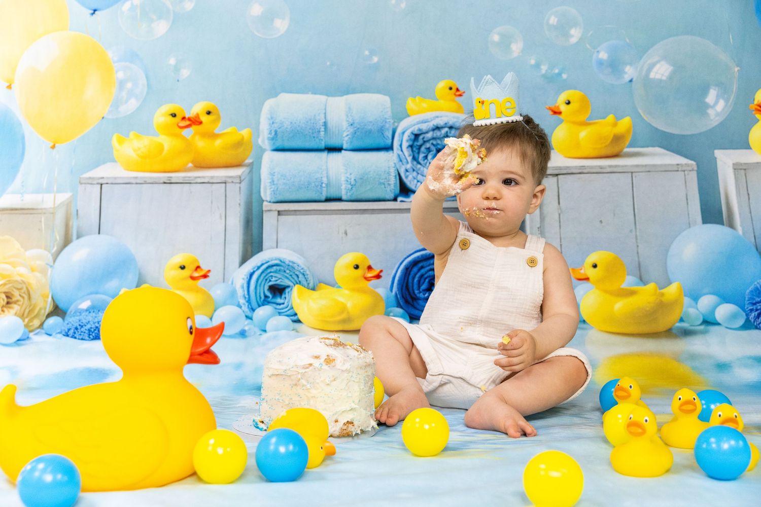 Cute toddler playing with rubber duck toys surrounded by yellow balloons during a fun first birthday photoshoot.