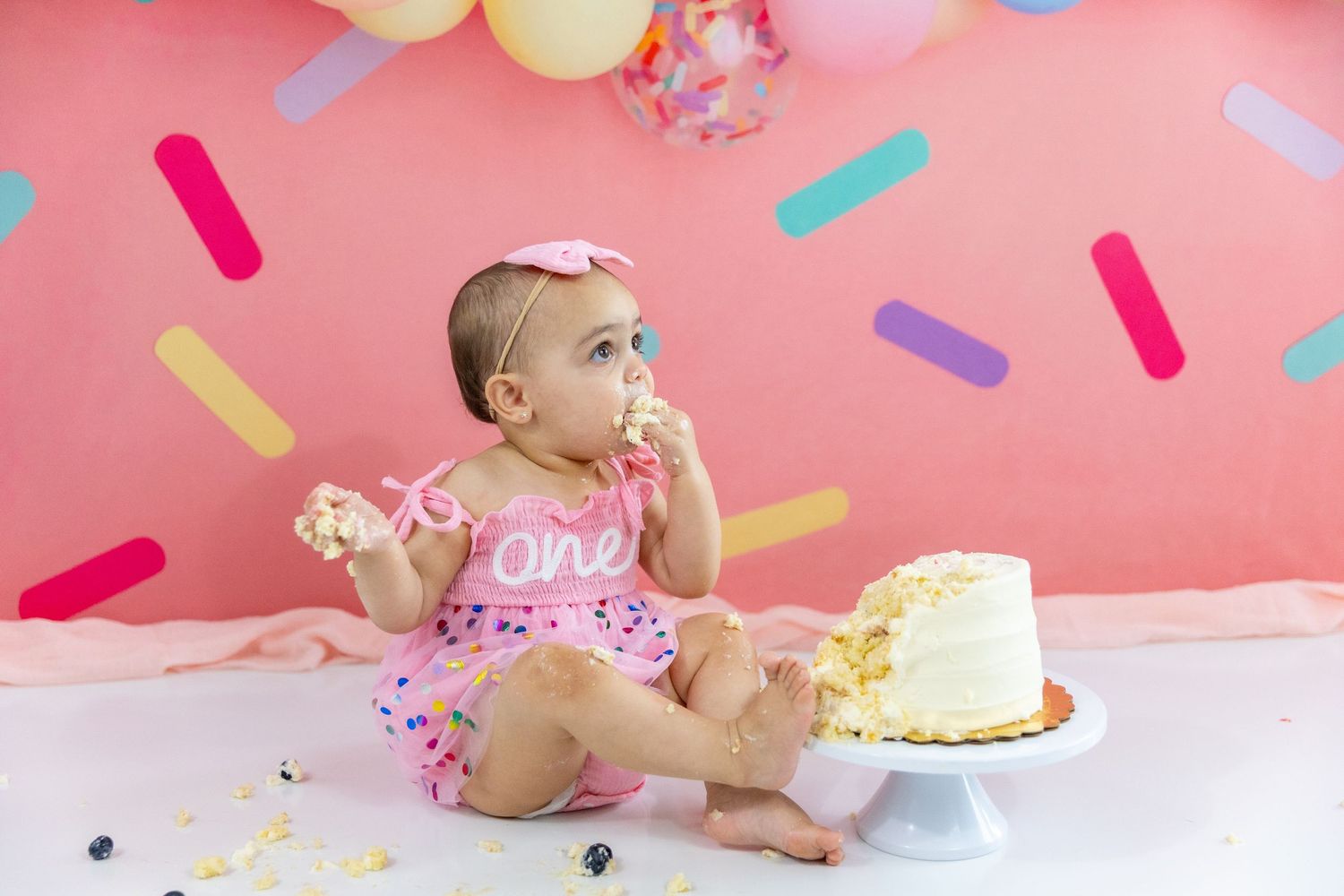 Baby girl in pink one outfit eating birthday cake during colorful confetti smash cake photo session.