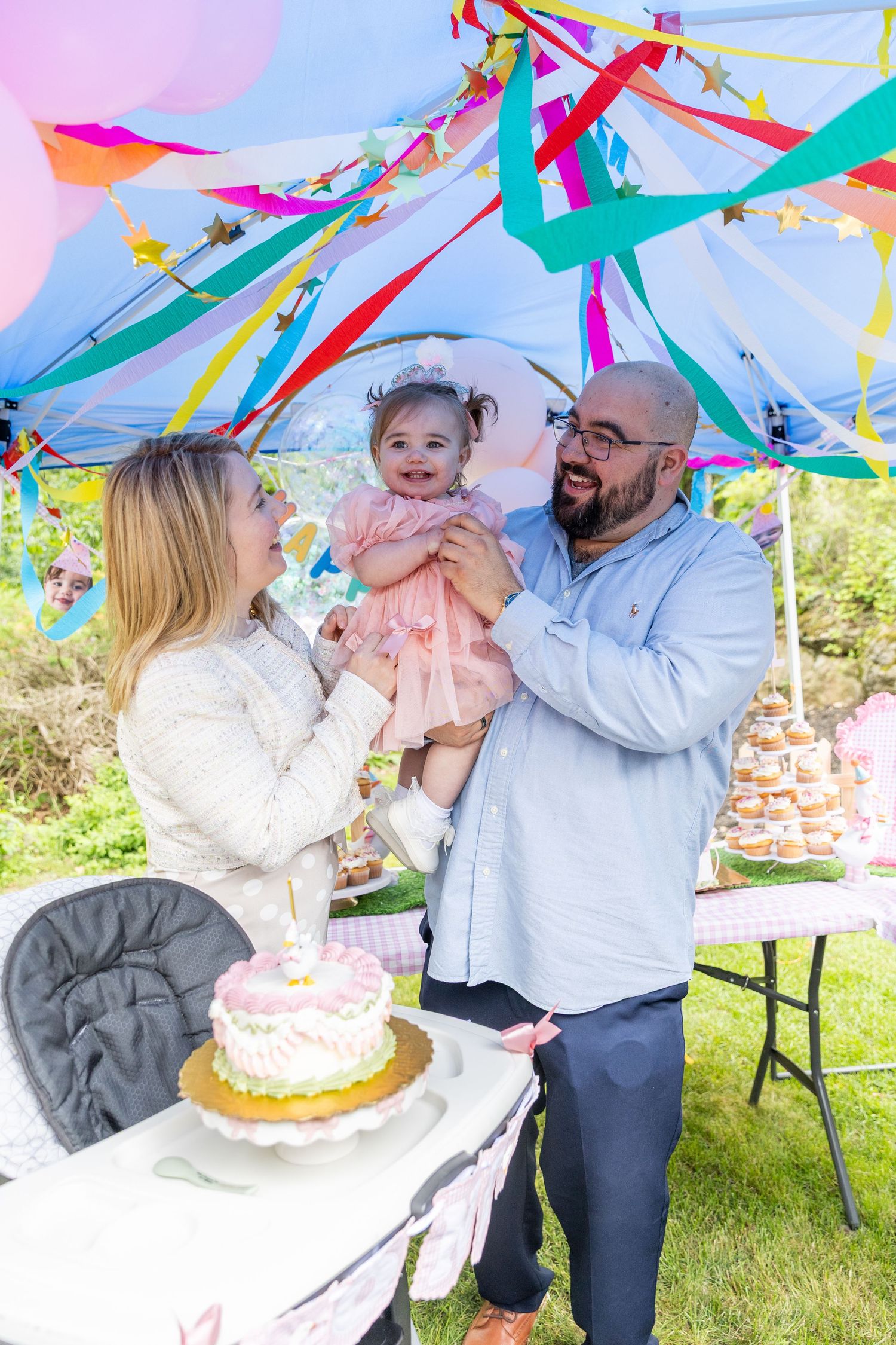 Happy parents holding their baby girl in a pink dress at a colorful outdoor first birthday party.