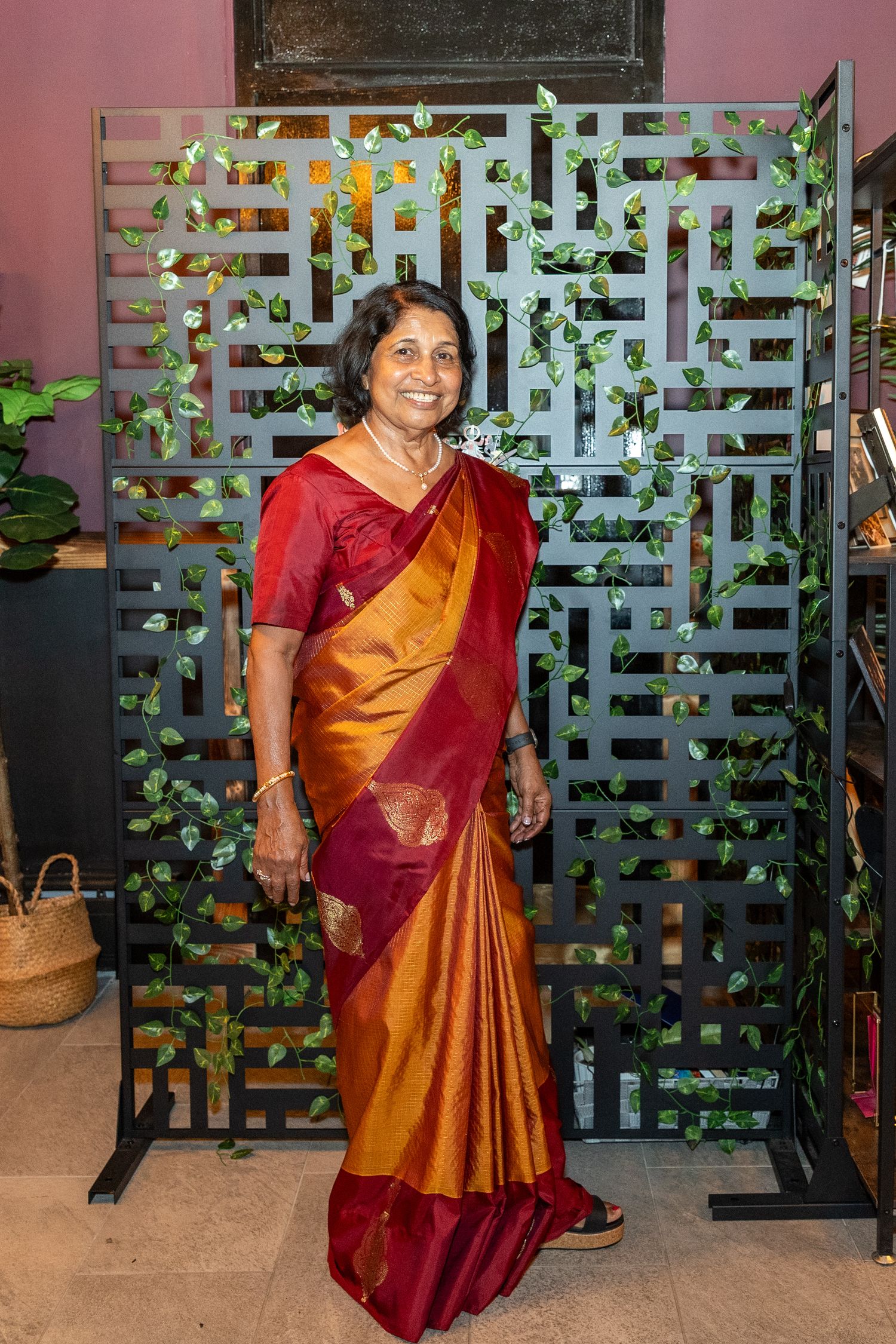Elegant elderly woman in a traditional red and gold silk saree posing in front of a decorative lattice backdrop.