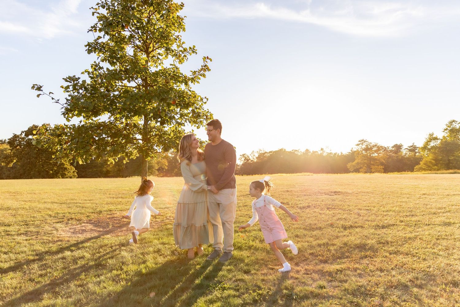 Family with two young girls playing in a sunlit green field with a large tree during golden hour.
