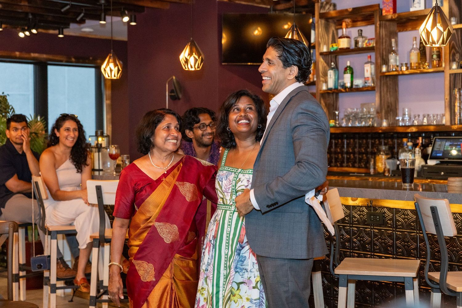Two women and a tall man laughing together near a bar at a festive indoor birthday party.
