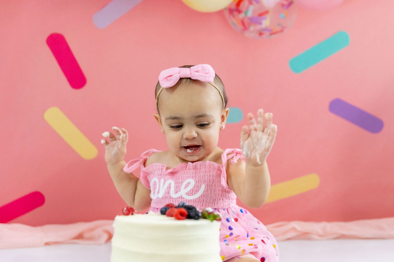 Baby girl in pink dress with bow headband clapping hands over white first birthday smash cake.