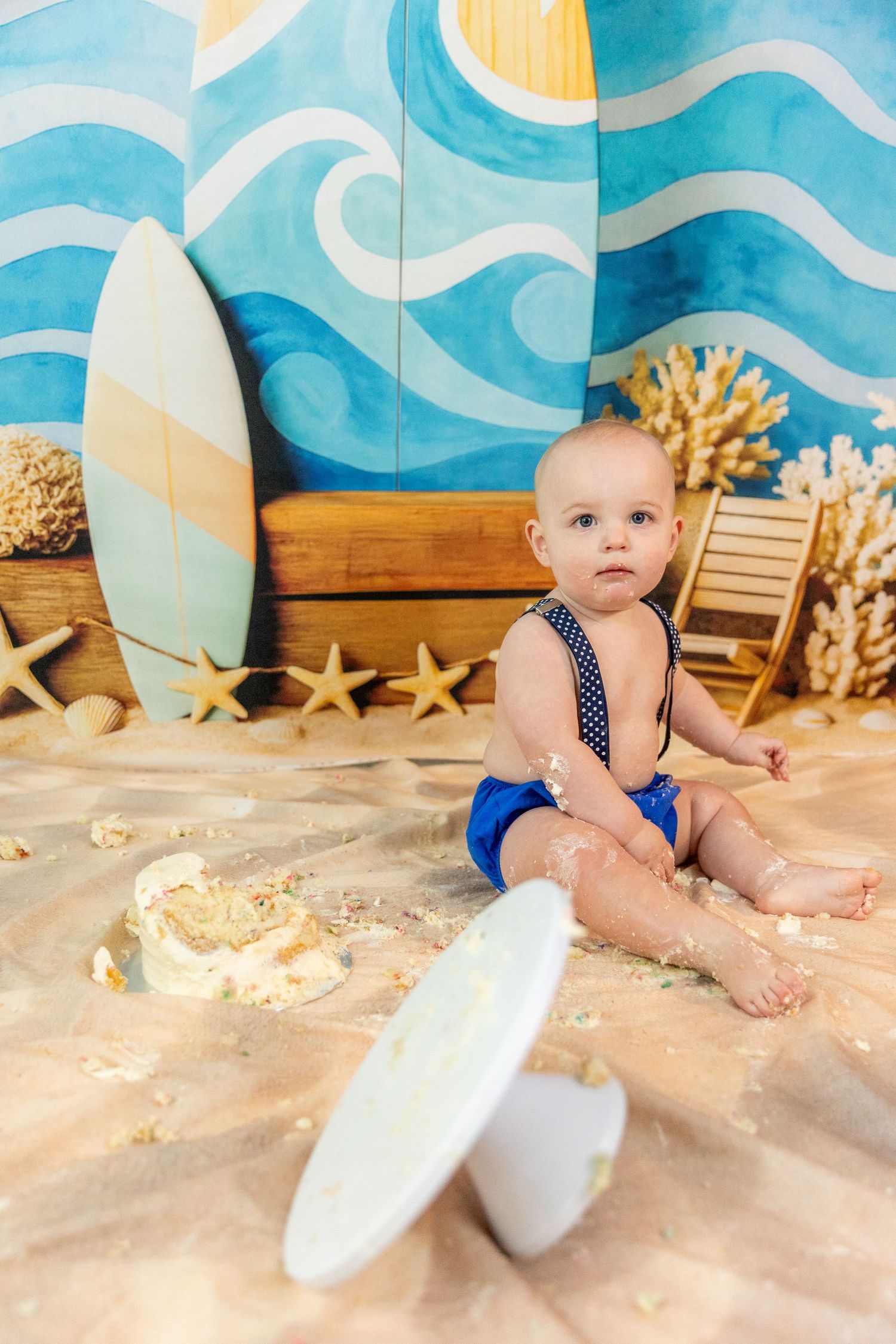 Baby boy in blue shorts sitting on sand surrounded by smashed cake with surfboard backdrop.