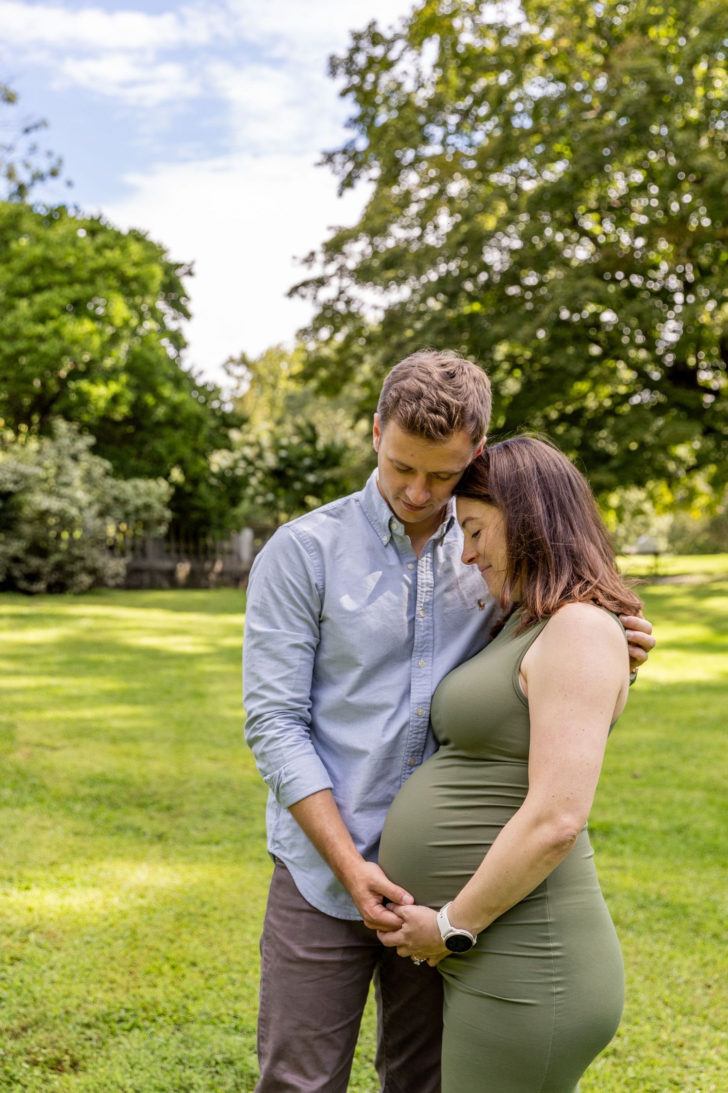 Couple embracing tenderly in green park, pregnant woman in olive dress during outdoor maternity shoot.
