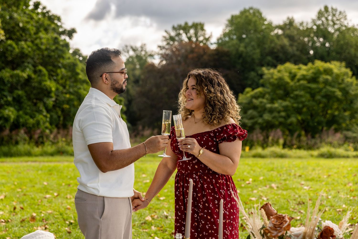 Happy couple toasting champagne glasses in green park during outdoor engagement celebration.
