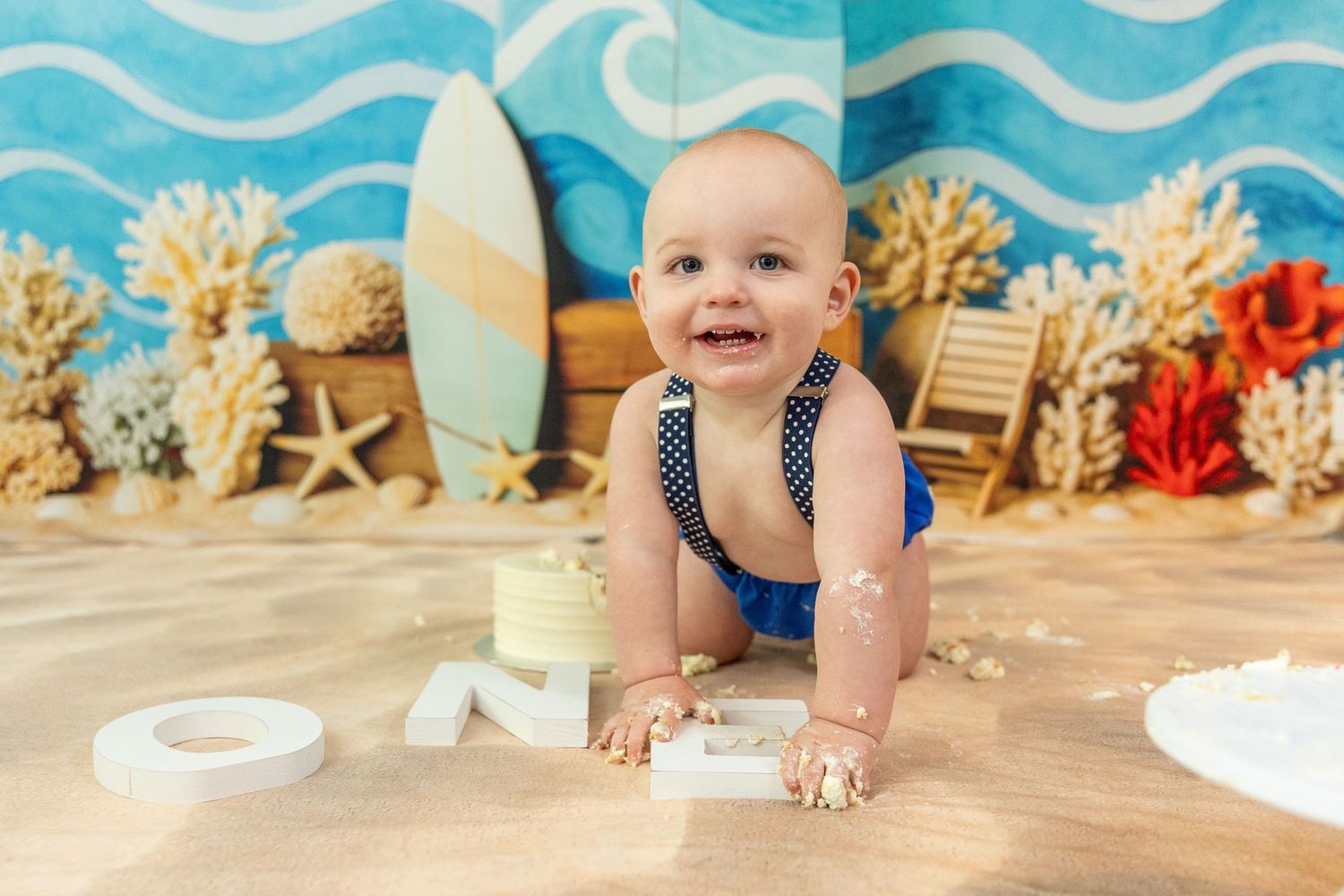 Baby boy crawling through cake smash mess on sandy beach-themed first birthday photo set.