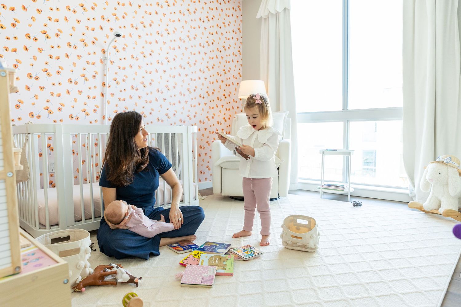 Mother sitting on nursery floor holding baby while toddler plays in decorated room with crib and toys.