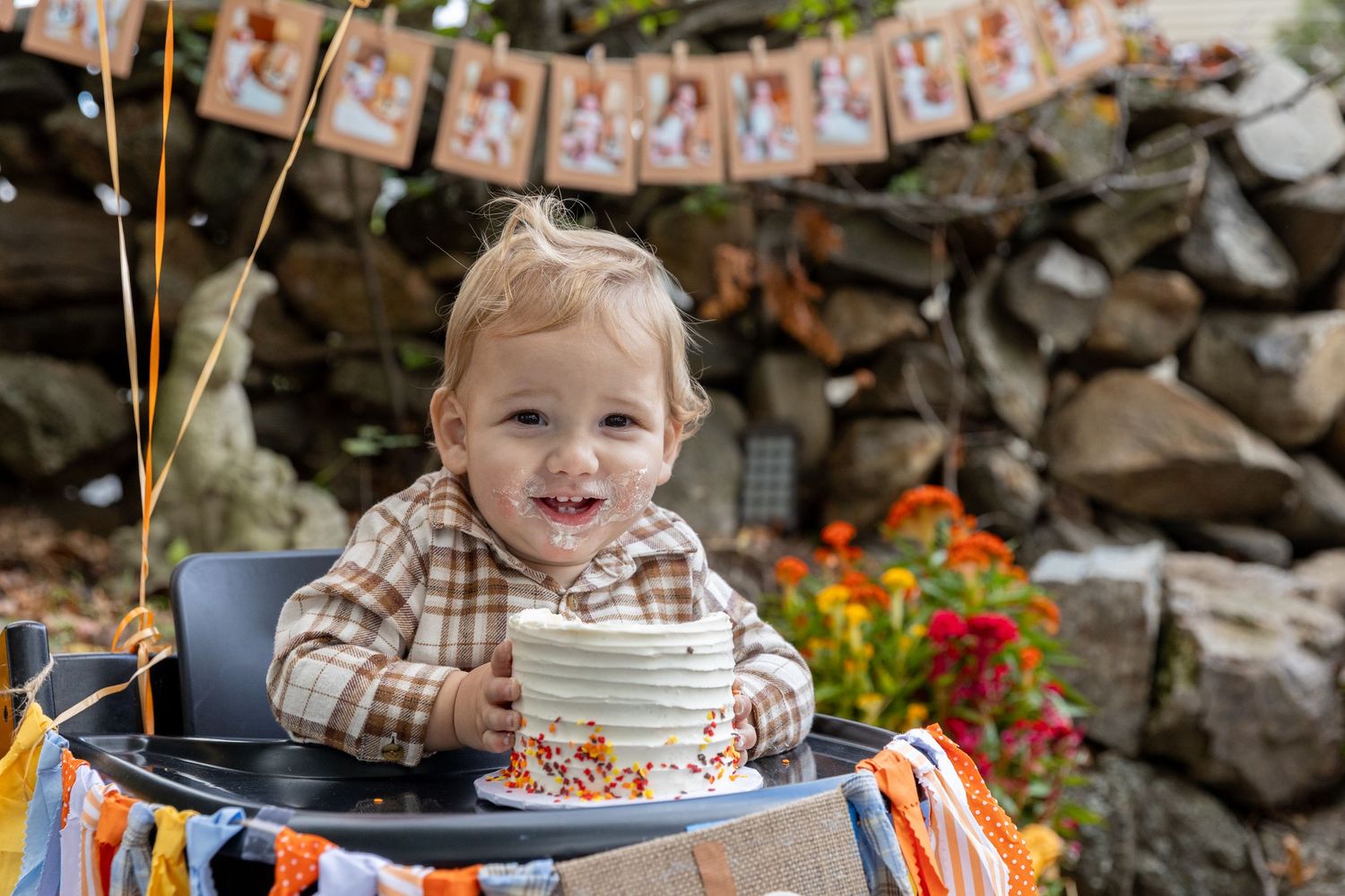 Smiling baby celebrating first birthday with cake in high chair at fall outdoor party.
