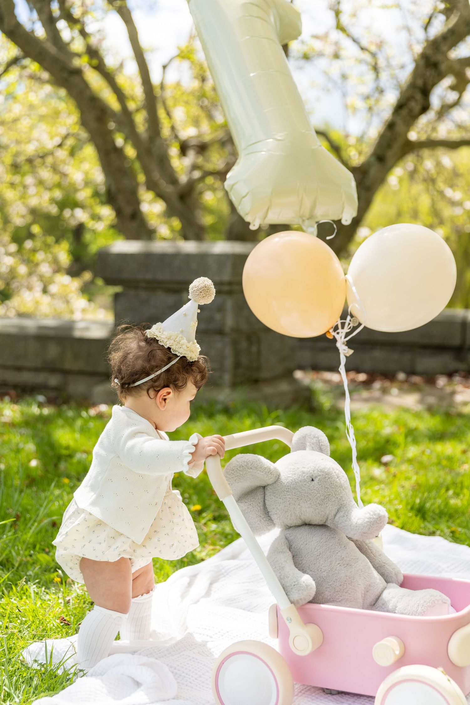 Baby girl in party hat standing at pink toy stroller with elephant and balloons in sunny garden.