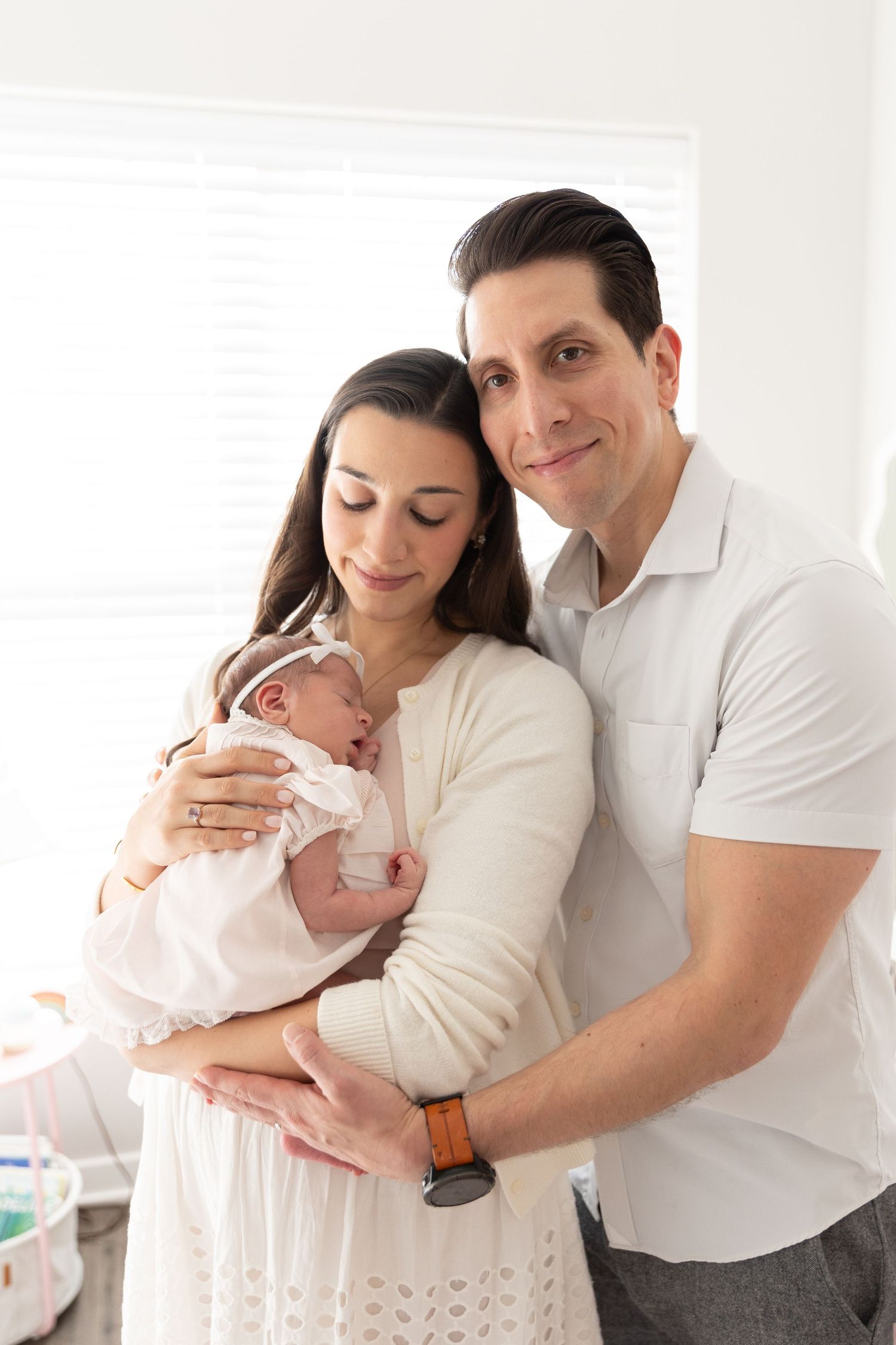 Smiling new parents proudly hold their newborn baby girl dressed in white near a bright window.