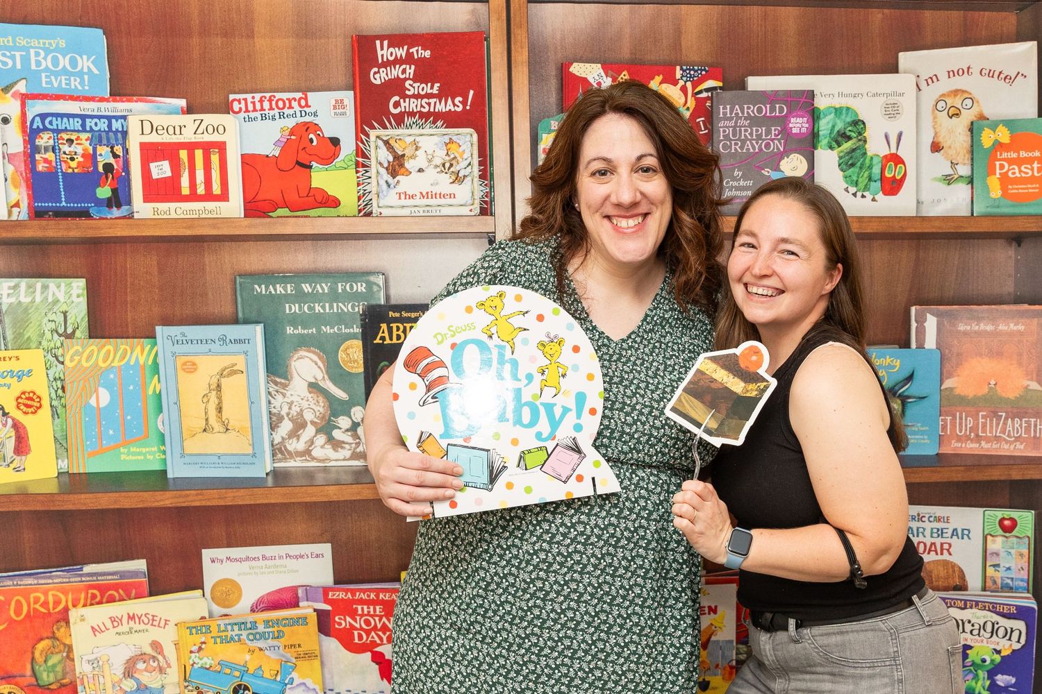 Two women smiling at a book-themed baby shower holding Oh Baby and cupcake photo props.